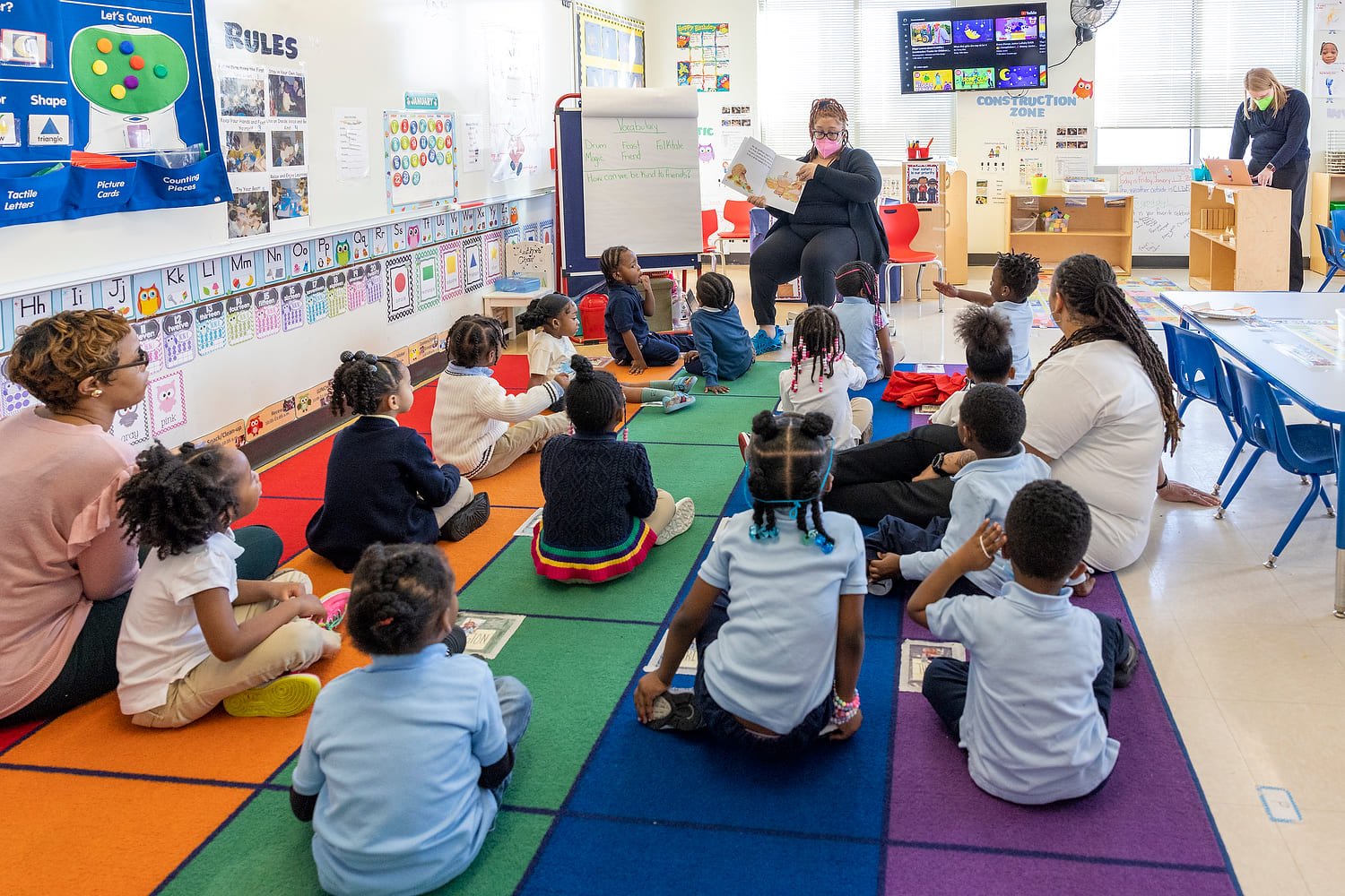 Photo of a classroom with students sitting on the floor and an adult reading them a book